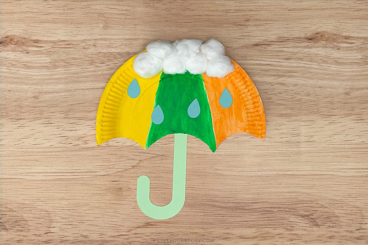 Finished umbrella paper plate craft with yellow, green, and orange canopy, raindrops, green handle, and white cotton balls for clouds