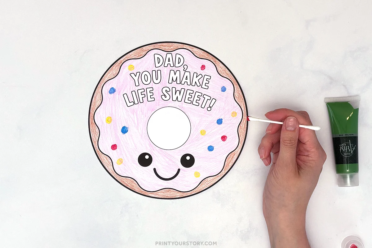 Hand painting a colored Father’s Day donut craft that says “Dad, you make life sweet!” using cotton swab