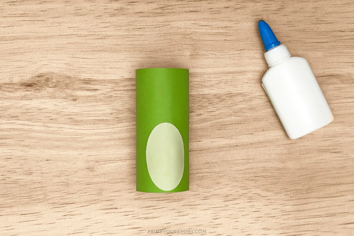 Green toilet paper roll frog body with a pale oval belly and glue bottle on a wooden table