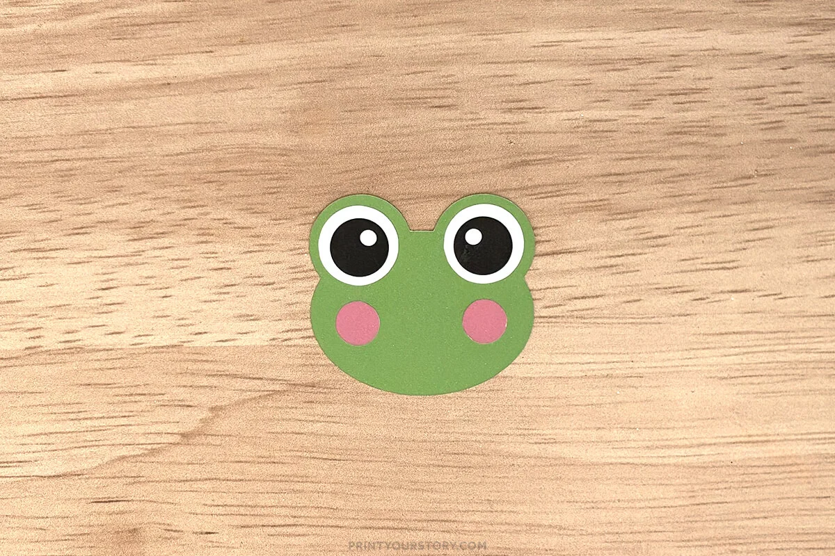 Green paper frog face with large eyes and pink cheeks on a wooden surface