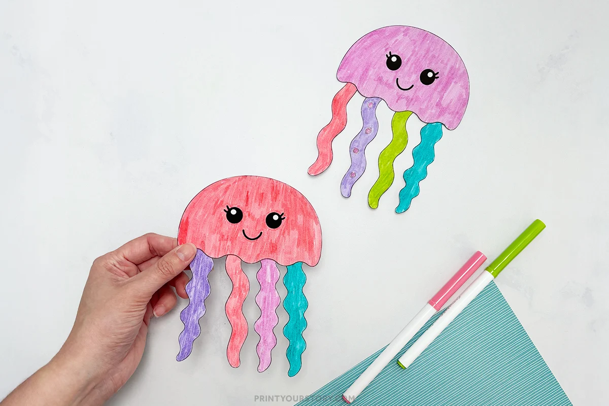 Hand holding a colorful paper jellyfish cut-and-paste craft with markers and another jellyfish on the table
