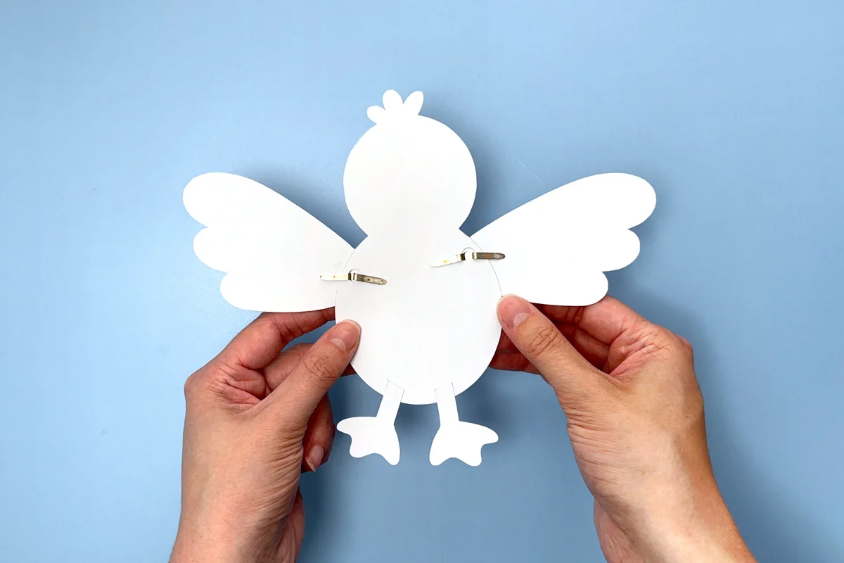 Hands holding a white paper seagull craft with wings attached by brass fasteners on a blue background