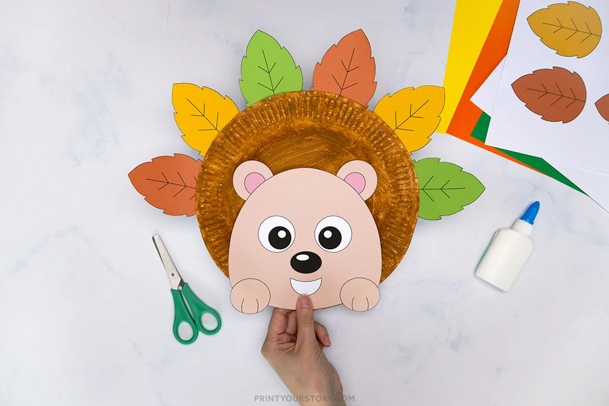 Paper plate hedgehog craft with colorful paper leaves, scissors, glue, and printable pieces on a table