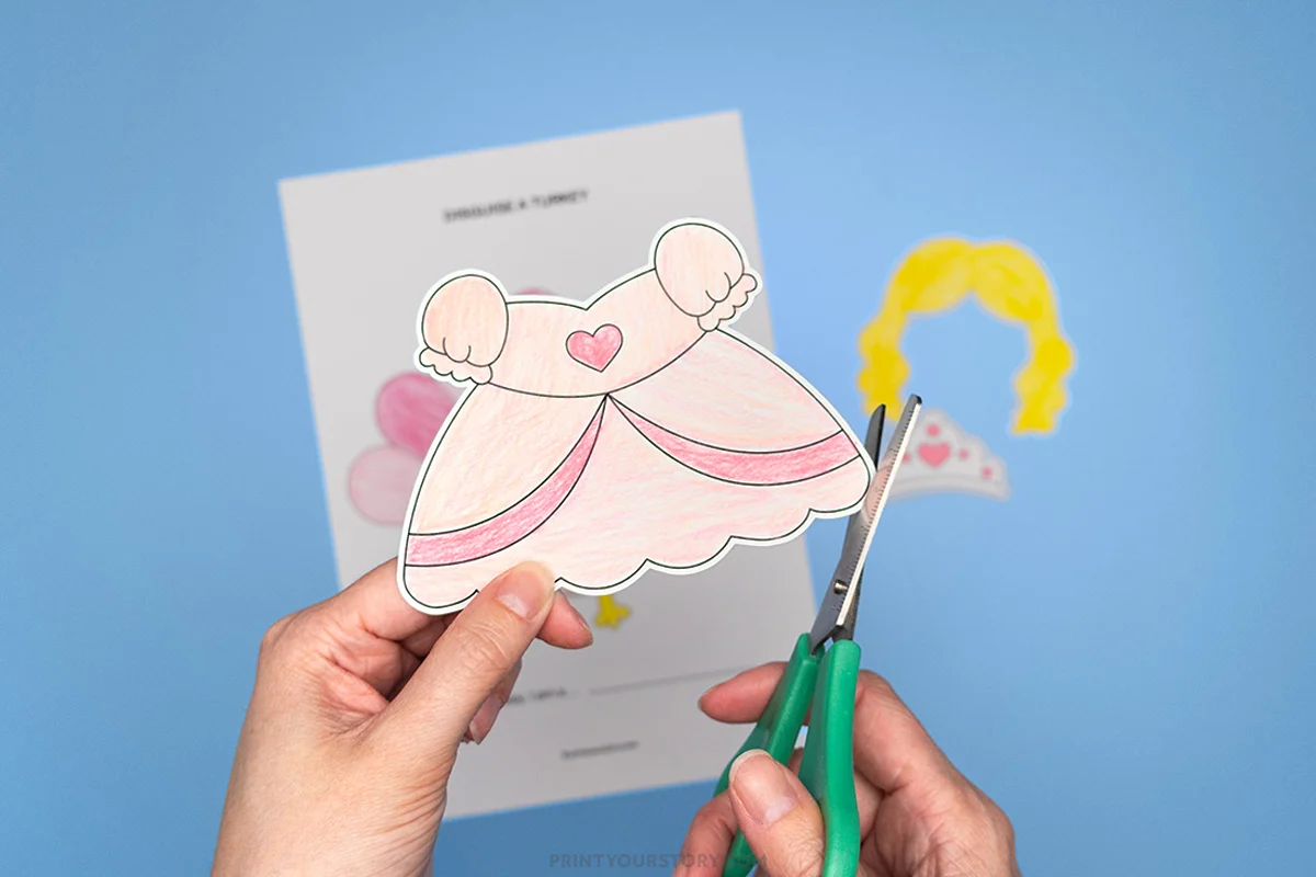 Hands cutting out a pink princess dress from a disguise a turkey printable template with wig and tiara pieces nearby