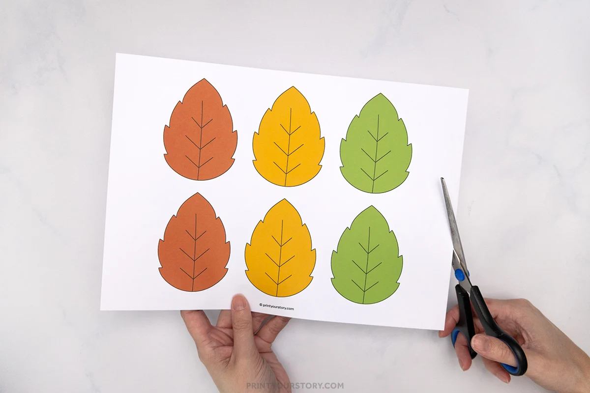 Hands holding scissors and a printed sheet of fall leaf cutouts for a paper plate hedgehog craft