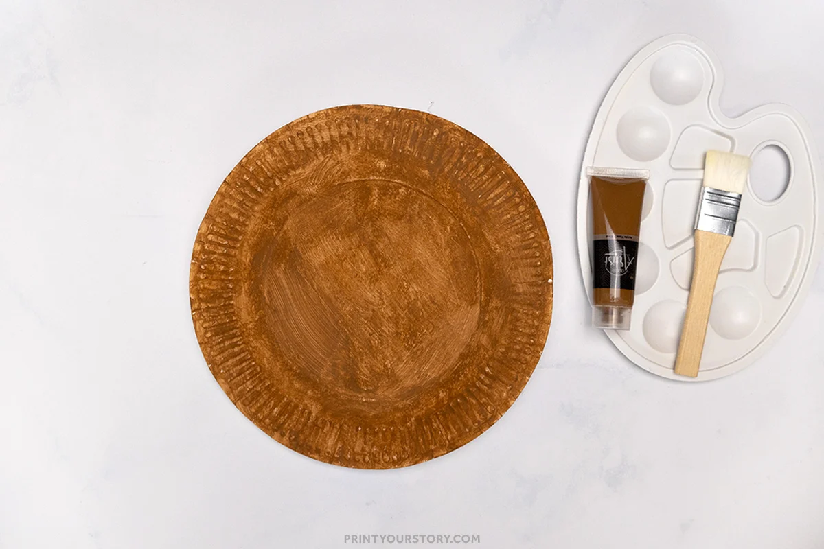 Brown-painted paper plate with brown paint tube, brush, and plastic palette for a hedgehog craft step