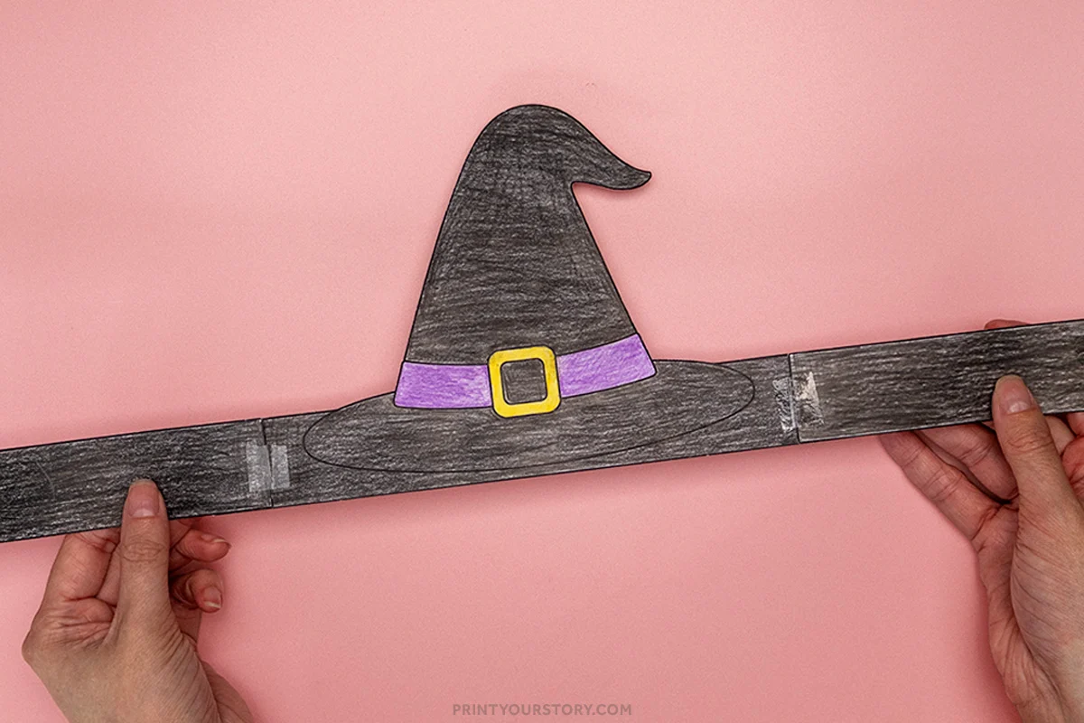 Hands holding a finished paper witch hat headband craft with a black hat, purple band, and yellow buckle