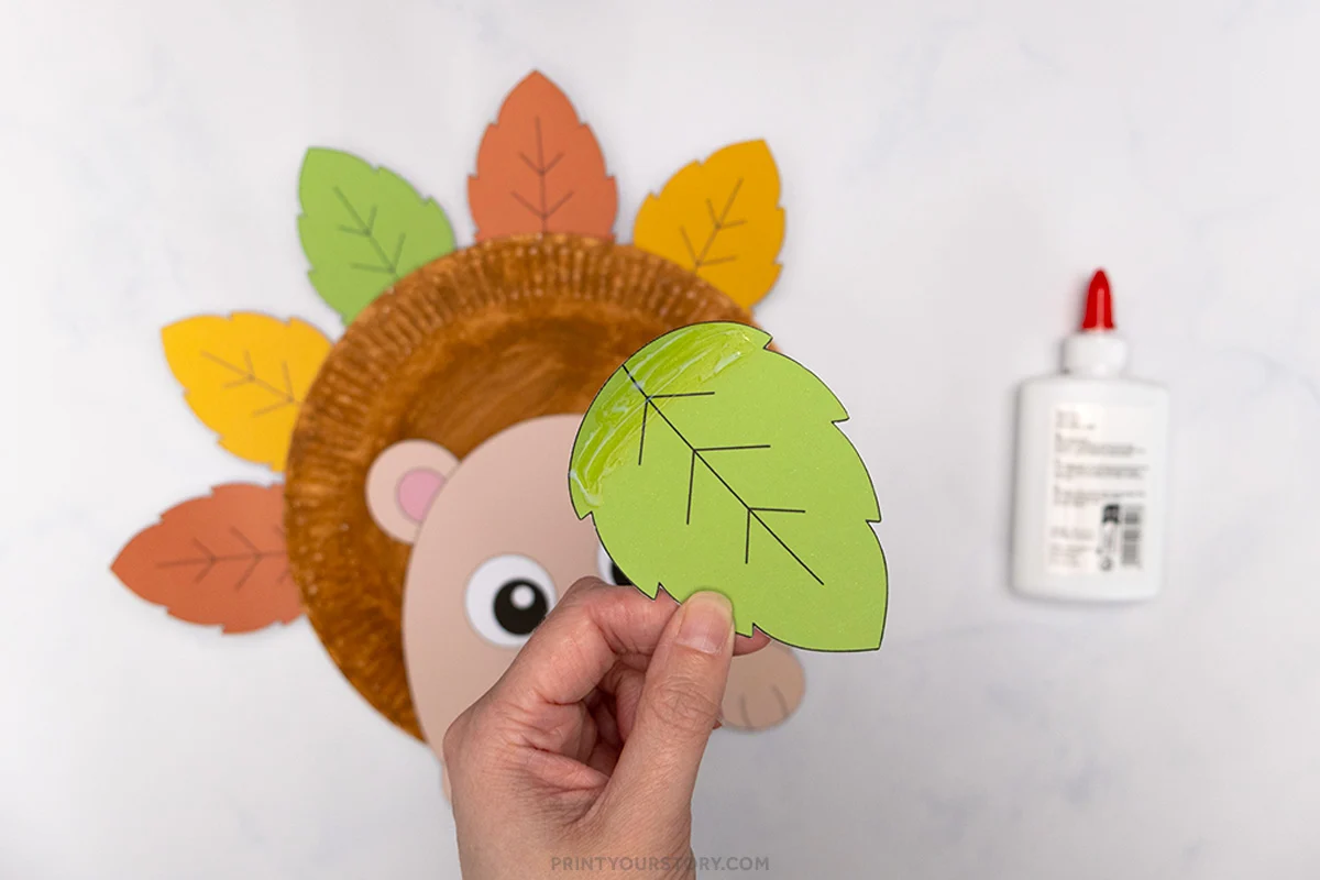 Hand gluing a green paper leaf onto a paper plate hedgehog craft, with colorful leaves and glue bottle nearby