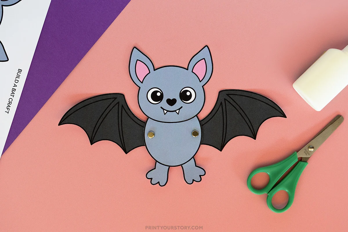 Printable build-a-bat Halloween craft with flapping black wings, beside scissors and a glue stick