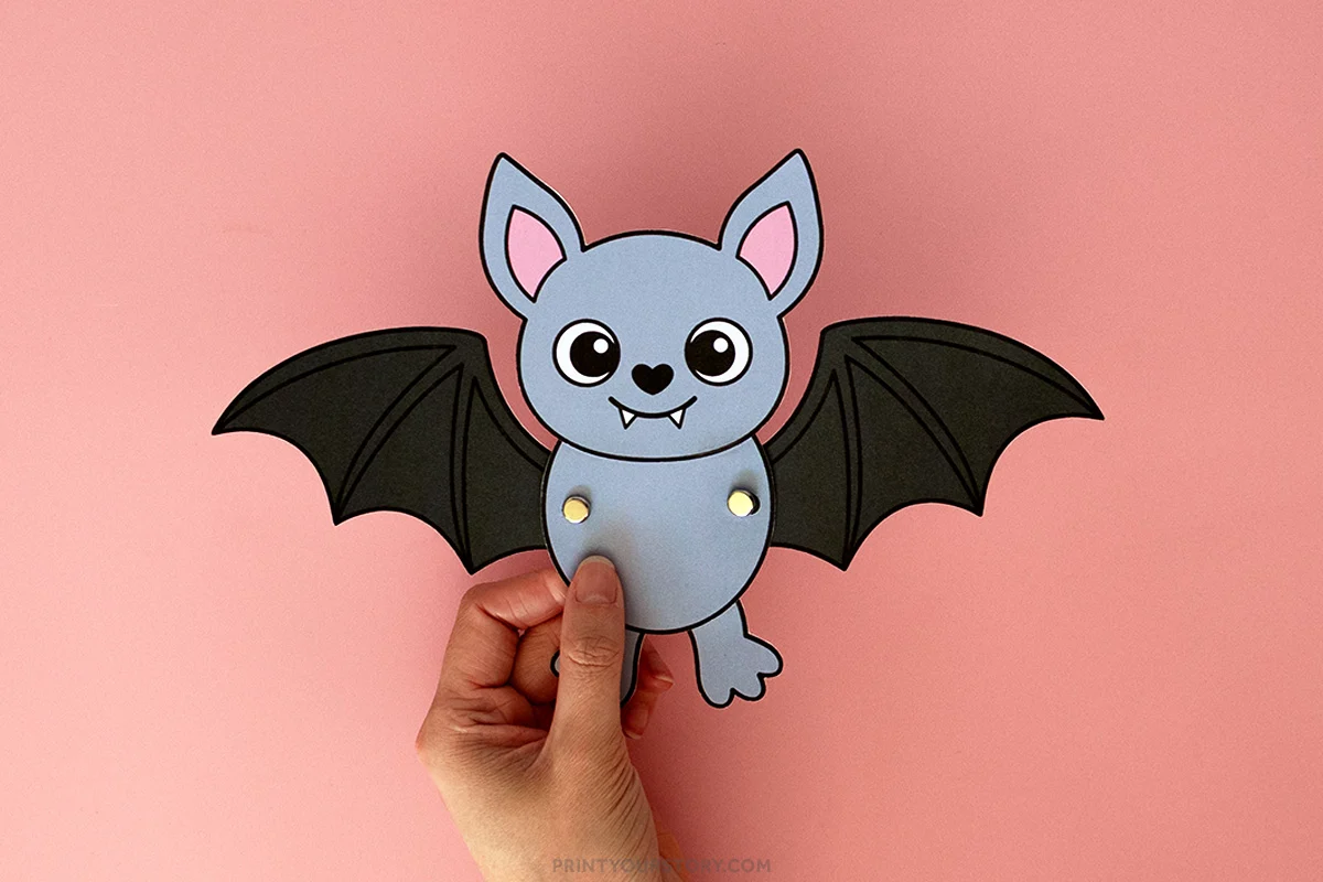 Hand holding a gray paper bat craft with flapping black wings on a pink background
