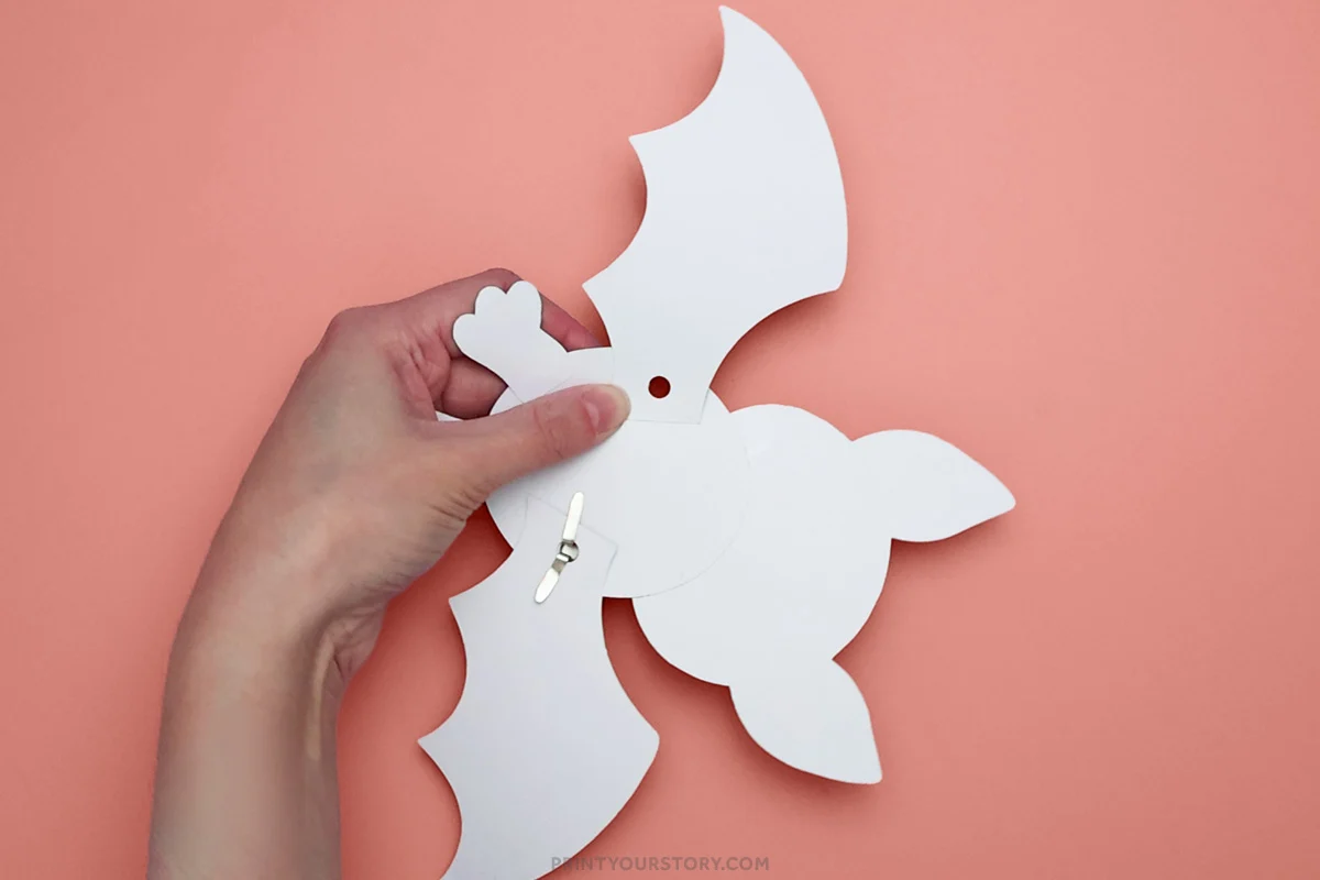 Hand holding the back of a white paper bat craft with a brad and wing mechanism on a pink background