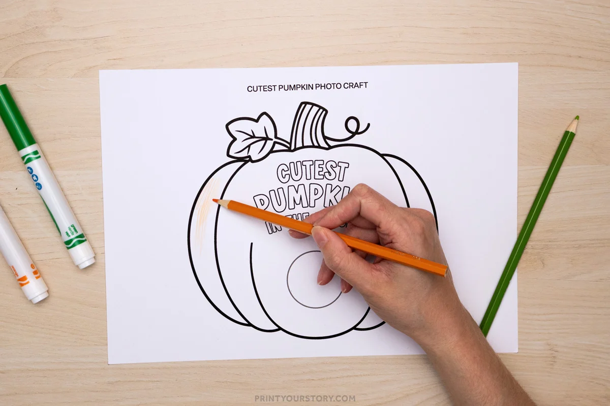 Hand coloring a pumpkin photo craft printable with an orange pencil on a table