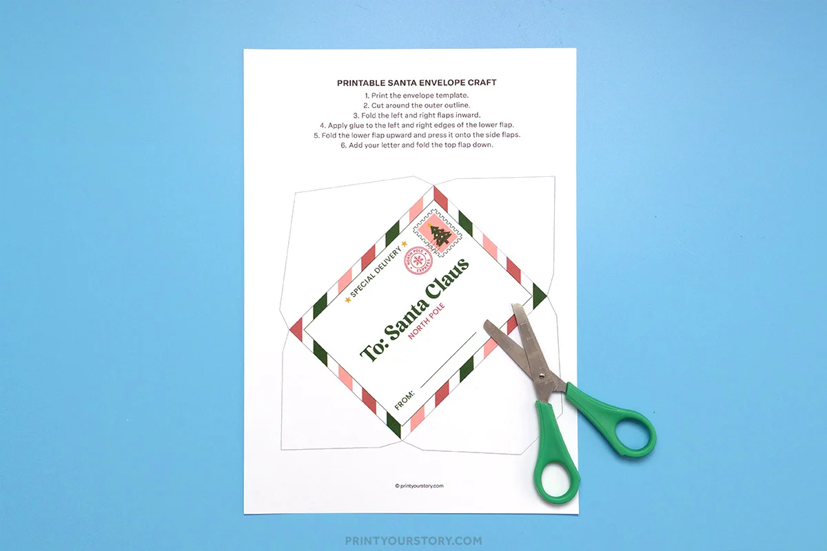 Printable Santa envelope template with cutting and folding instructions and green-handled scissors on blue background