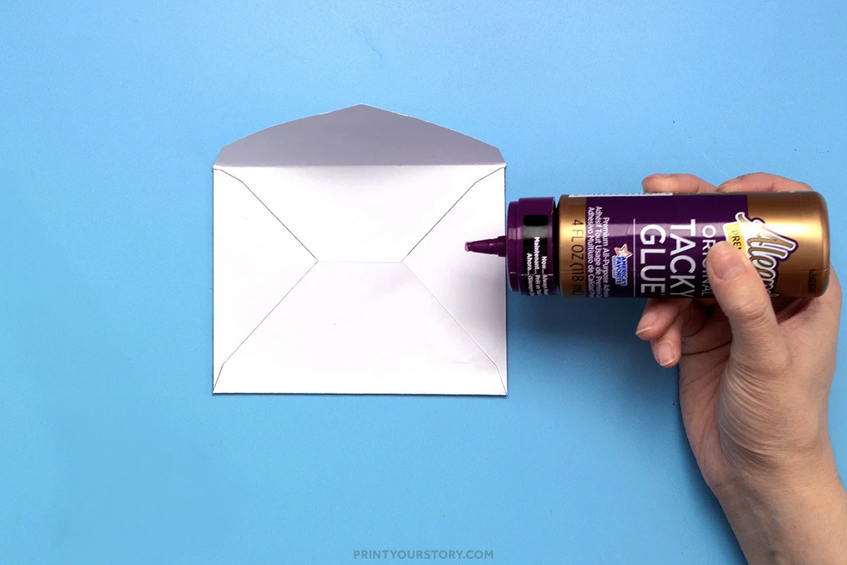 Applying tacky glue to the back center of a folded printable envelope on a blue background